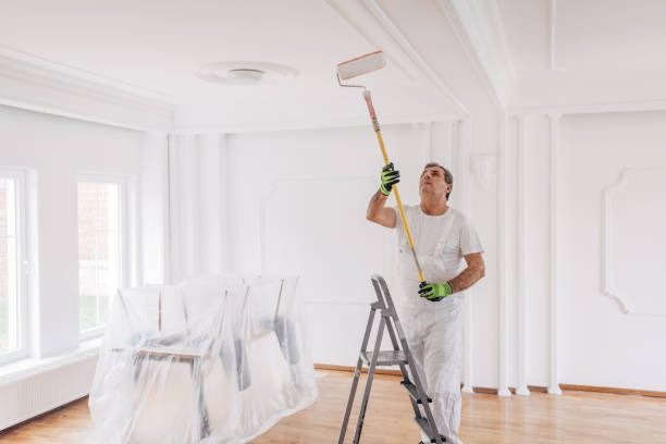 Painter in white overalls rolling a ceiling from a stepladder in an empty room