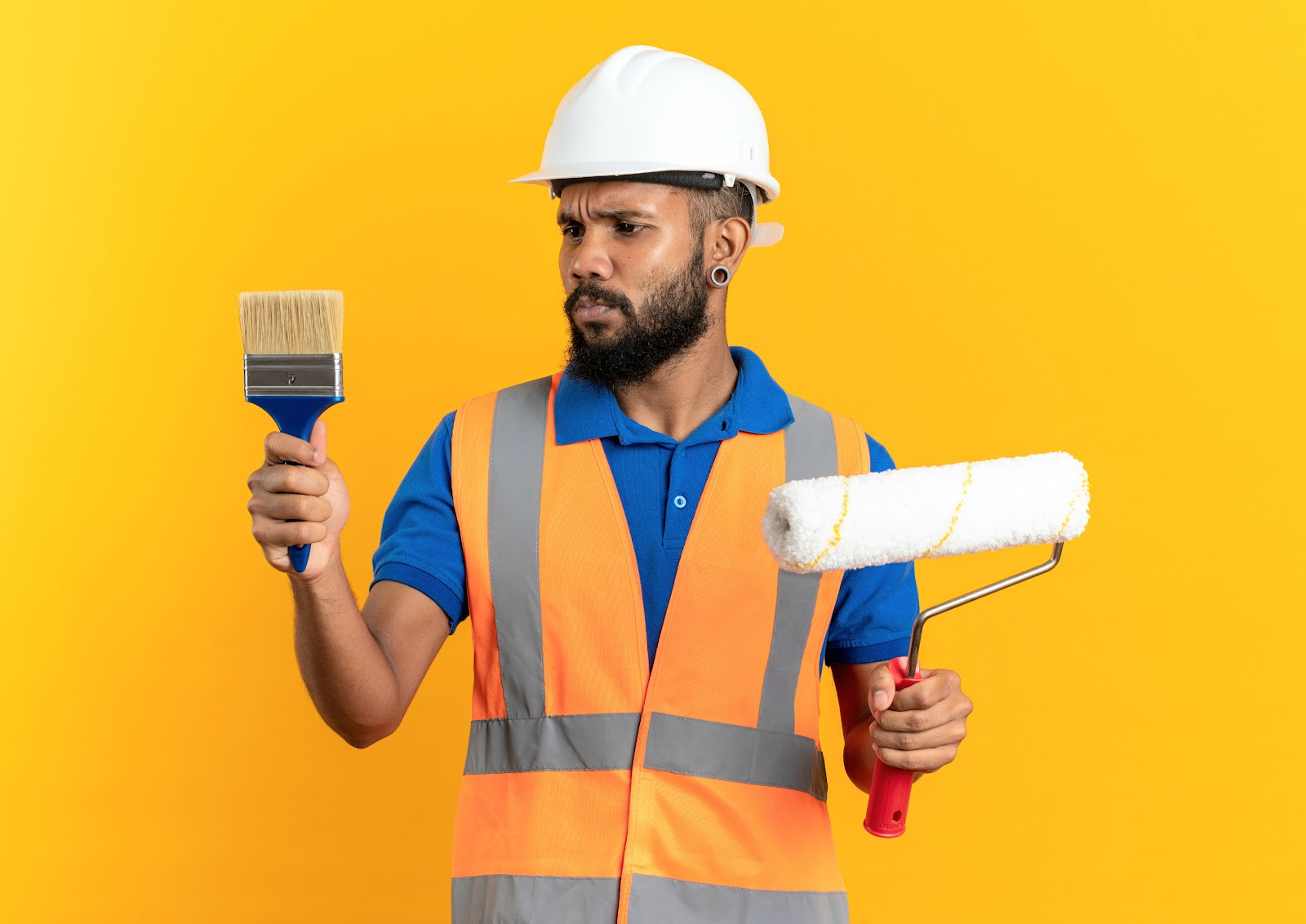 Painter in a hard hat and safety vest holding a brush in one hand and a roller in the other