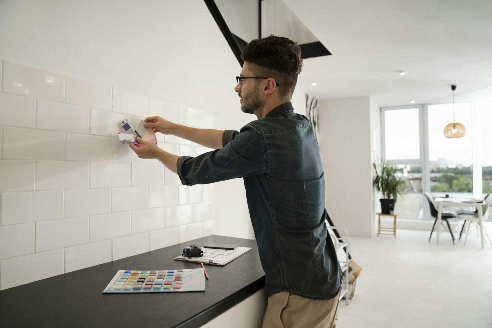 A man holding a color swatch against a white tiled wall in a bright apartment