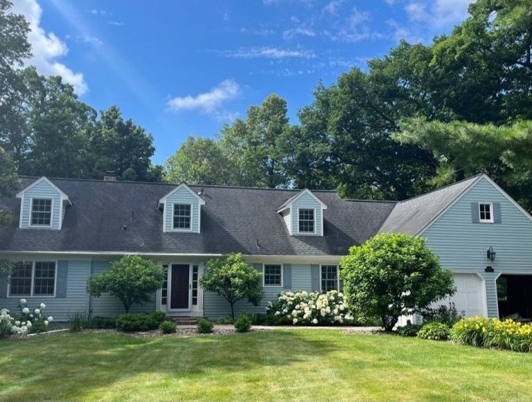 Cape Cod house in pale blue with dormer windows and white trim, behind flowering shrubs